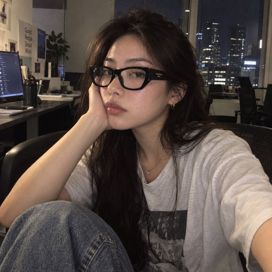 A woman with long dark hair and Balenciaga BB0372O 001 Black Butterfly Eyeglasses posing in an office setting at night, wearing a light gray graphic t-shirt and blue jeans.