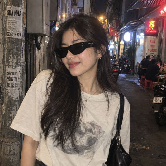 A woman wearing Balenciaga BB0388S 001 Black Rectangular Sunglasses, a beige graphic t-shirt, and a black shoulder bag, standing in a nighttime street setting.