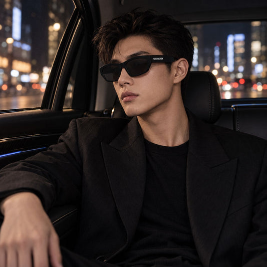 A man wearing Balenciaga BB0448SK 001 Black Rectangular Sunglasses and a black blazer sits in the back of a car at night with blurred city lights visible through the window.
