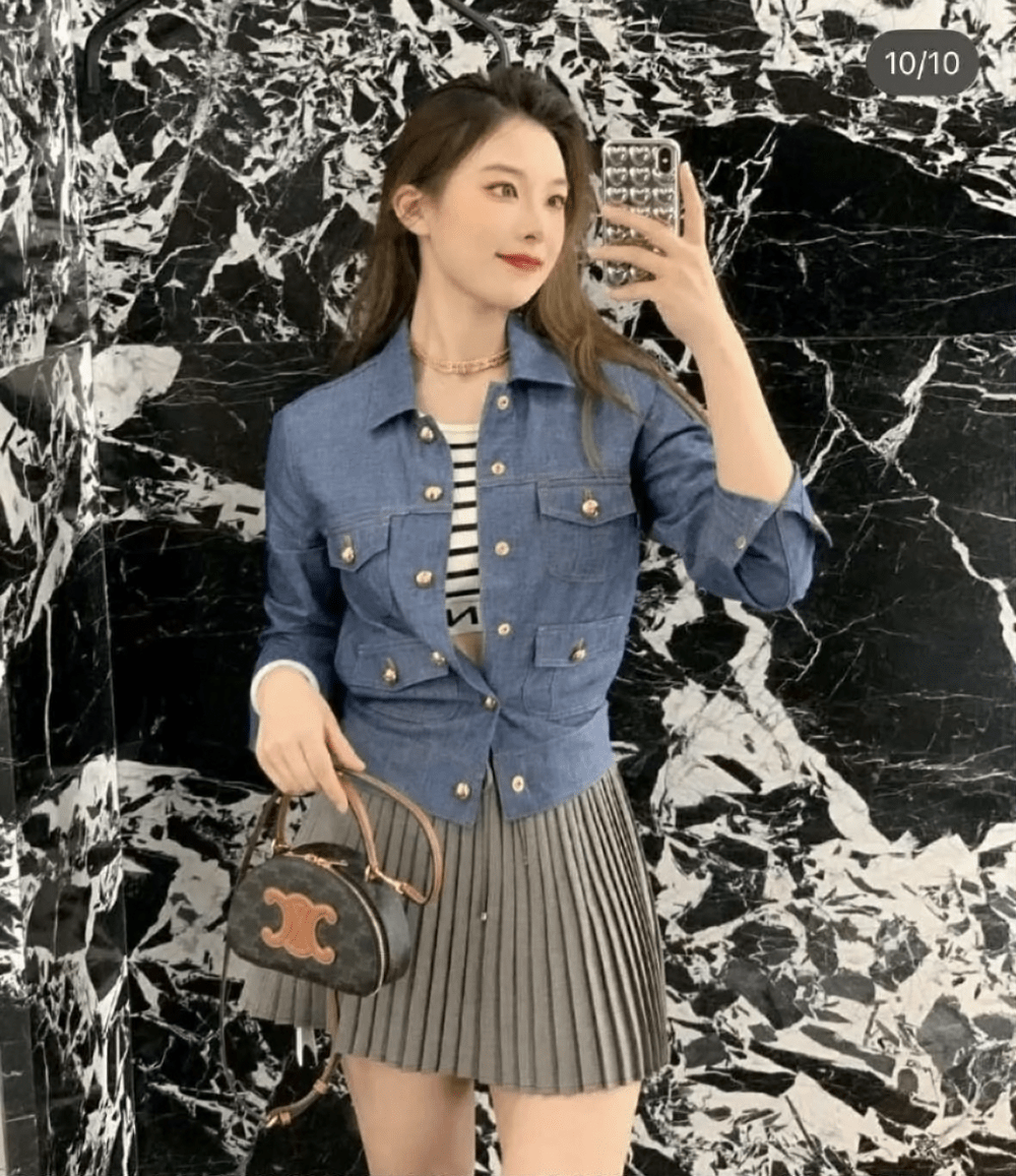 Woman taking a selfie wearing a blue denim jacket and pleated skirt and holding a Celine Half Moon Case In Triomphe Canvas & Calfskin 10K642ECM.04LU against a marble wall.