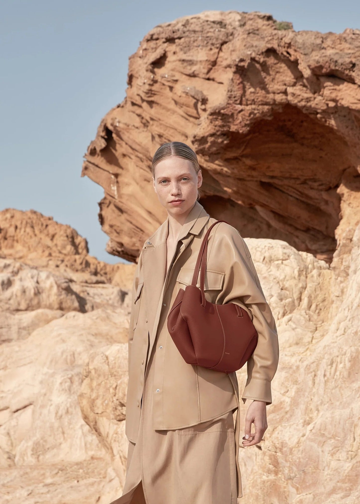 Woman in beige outfit with brown bag standing in front of rock formations