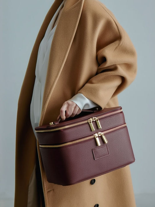 Person wearing a brown coat holding a Etoile Duo Vanity Case Ginger Burgundy with gold zippers against a light gray background