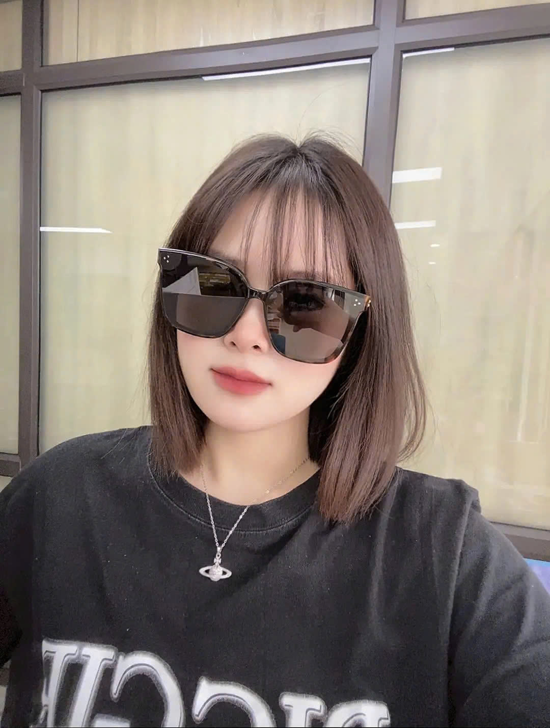 woman wearing Gentle Monster (GM) New Her T1 and a black shirt with white text, indoors.