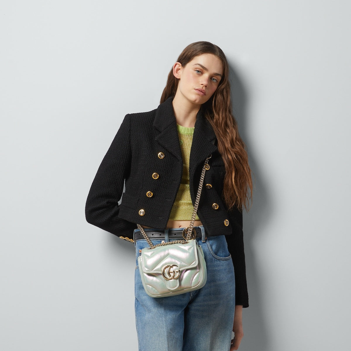 Woman wearing a black jacket with gold buttons, holding a Gucci GG Marmont Iridescent Mini Crossbody Bag in Green 739682 AAC2U 3404 against a light gray background