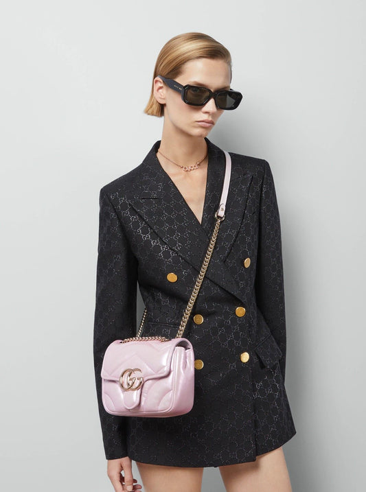 Woman wearing a black textured blazer with gold buttons and carrying a Gucci GG Marmont Iridescent Mini Crossbody Bag in Pink 739682 AAC2U 5826 on a light gray background