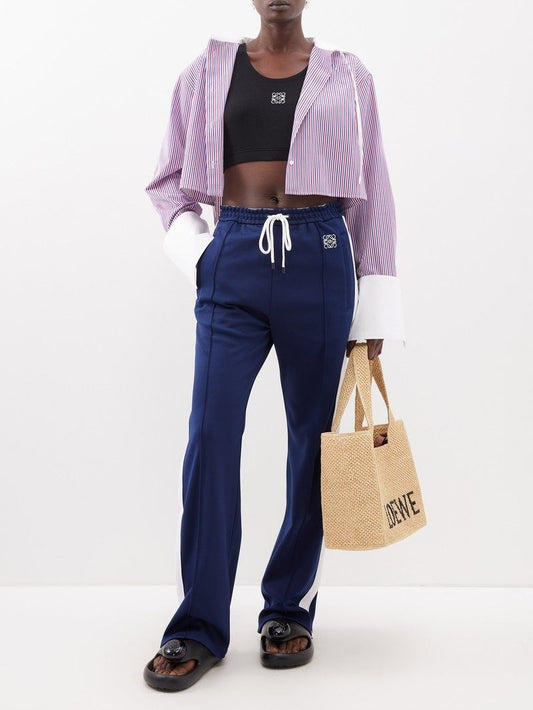 Person wearing a purple cropped hoodie, black top, and blue pants, holding a straw bag with Loewe Paula Ibiza Foam Thong Slide Black L616S03X02 1100.