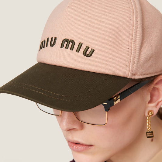 Person wearing Miu Miu Orchid Pink Military Drill Baseball Cap 5HC179 ACQ1 F03RG, gold glasses, and earrings on a light gray background.