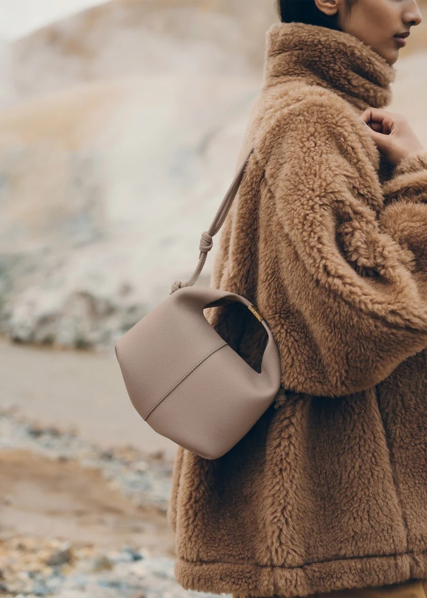Person wearing a fluffy brown coat holding a Polene Béri Edition Textured Taupe BHB0014MGRBR007 with a blurred natural background