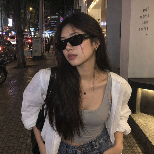 A woman wearing Saint Laurent SL 740 005, a white button-up shirt, a gray ribbed tank top, and blue jeans, standing on a city sidewalk at night.