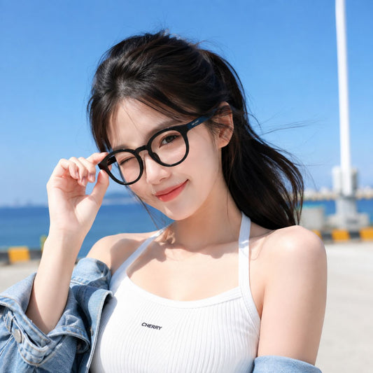 A woman outdoors by the water wearing Saint Laurent SL 907/F 001 Black Round Eyeglasses, a white ribbed tank top, and a light blue denim jacket. The background shows a bright blue sky and sea.