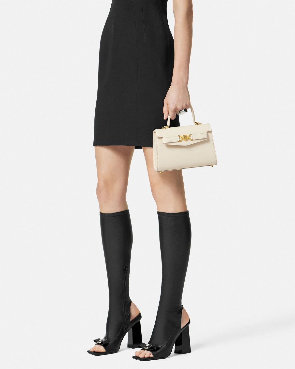 Person wearing a black dress, black knee-high boots, and holding a Versace Medusa '95 Small Handbag Cream 1013824 1A10795 1KD7V on a white background