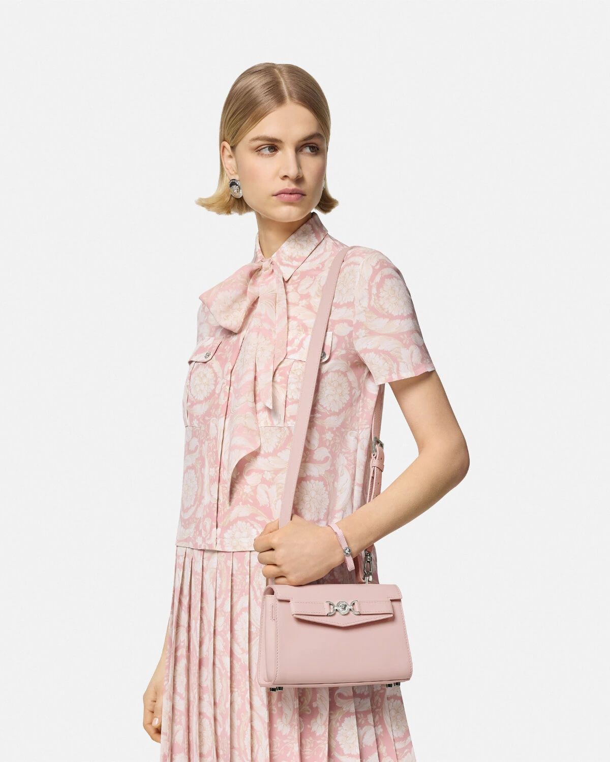 Woman wearing a pink patterned dress holding a matching Versace Medusa '95 Small Handbag Light Pink 1013824 1A10795 1PT2P on a light gray background