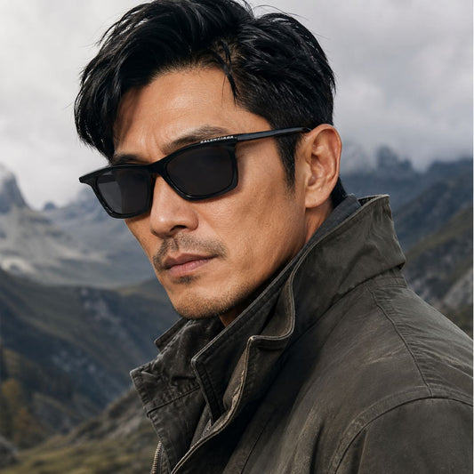 A man wearing Balenciaga BB0099S-001 Black Squared Sunglasses and a dark olive jacket, photographed outdoors against a cloudy mountain landscape. The sunglasses appear to be the featured product.