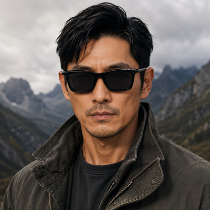 A man wearing Balenciaga BB0099S-001 Black Squared Sunglasses and a dark jacket, photographed outdoors against a cloudy mountain landscape.