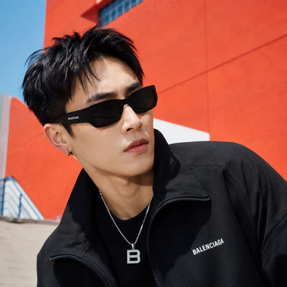 A man wearing Balenciaga BB0331SK-001 Black Squared Sunglasses and a black Balenciaga zip-up jacket, standing outdoors against a bright orange building wall.