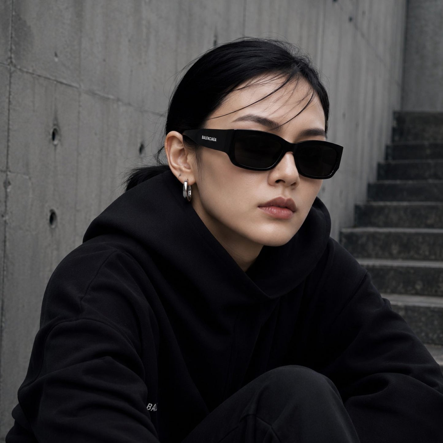 A woman wearing Balenciaga BB0331SK-001 Black Squared Sunglasses with a small white brand logo on the temple, paired with a black hoodie, sitting against a gray concrete wall and staircase.