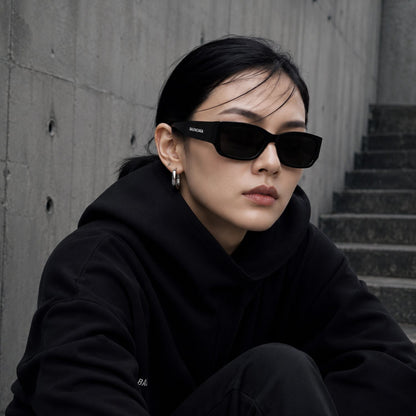 A woman wearing Balenciaga BB0331SK-001 Black Squared Sunglasses with a small white brand logo on the temple, paired with a black hoodie, sitting against a gray concrete wall and staircase.