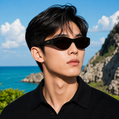 A man wearing Balenciaga BB0367S Round Sunglasses and a black polo shirt, photographed outdoors against a sunny coastal background with blue ocean, sky, and rocky cliffs.