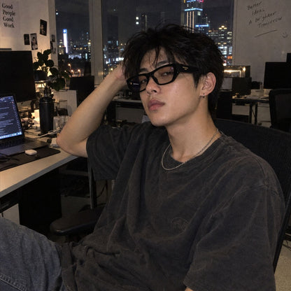 A young man with dark hair and Balenciaga BB0372O 001 Black Butterfly Eyeglasses sits in a dimly lit office workspace, wearing a faded black t-shirt and blue jeans. The background shows a city skyline at night through a window, computer desks, and office equipment.