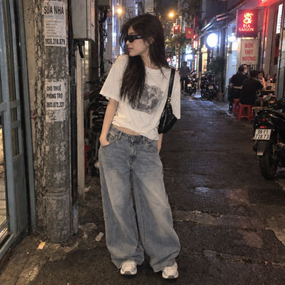 A woman stands on a nighttime street wearing a Balenciaga BB0388S 001 Black Rectangular Sunglasses in white graphic T-shirt, very wide-leg light-wash jeans, white sneakers, and a black shoulder bag. The setting is a real urban alley with storefronts, signs, and parked motorcycles, so this is Lifestyle rather than Product/Packaging.