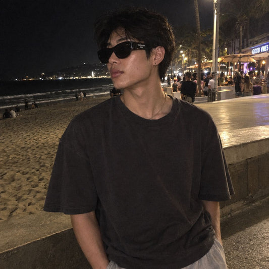A man wearing a dark charcoal gray oversized t-shirt and Balenciaga BB0389SK Black Rectangular Sunglasses stands on a nighttime beach boardwalk with a beach and city lights in the background.