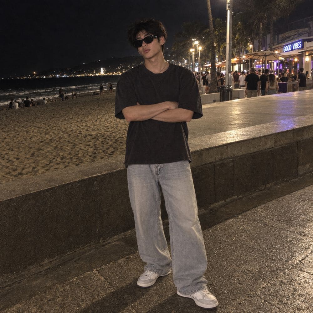 A man wearing a Balenciaga BB0389SK Black Rectangular Sunglasses in black oversized t-shirt, light-wash wide-leg jeans, and white sneakers stands on a nighttime beach boardwalk with his arms crossed.