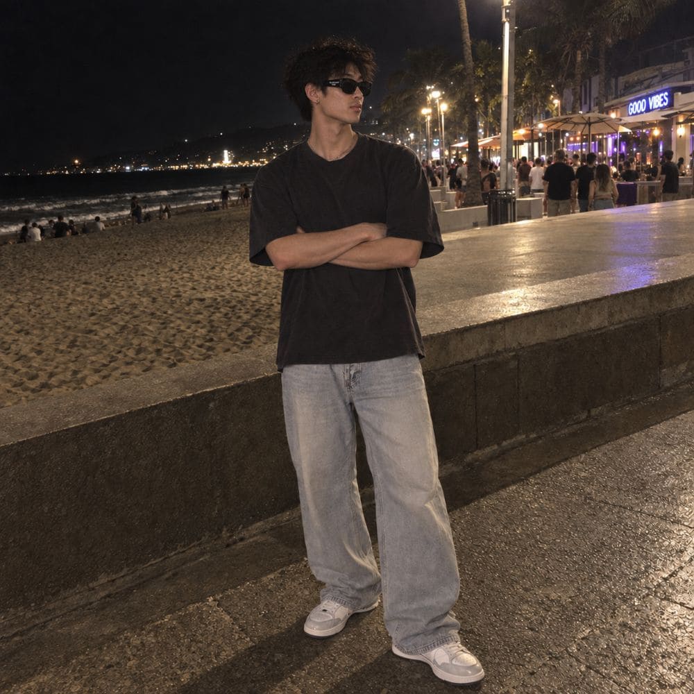 A man wearing a Balenciaga BB0389SK Black Rectangular Sunglasses in black oversized t-shirt, light-wash wide-leg jeans, and white sneakers stands on a nighttime beach boardwalk with his arms crossed.