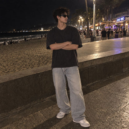 A man wearing a Balenciaga BB0389SK Black Rectangular Sunglasses in black oversized t-shirt, light-wash wide-leg jeans, and white sneakers stands on a nighttime beach boardwalk with his arms crossed.