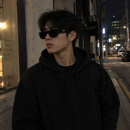 A man wearing a black hoodie and black Balenciaga BB0390SK 001 Black Square Sunglasses standing on a city street at night.
