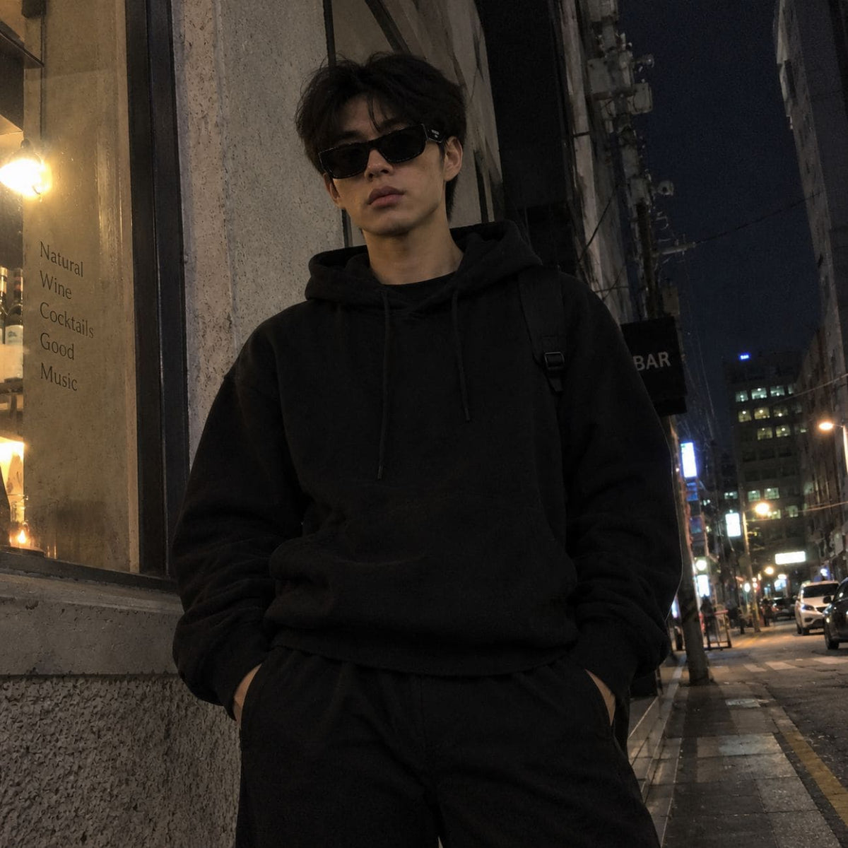 A man wearing a black hoodie, black sweatpants, and Balenciaga BB0390SK 001 Black Square Sunglasses stands on a city street at night.