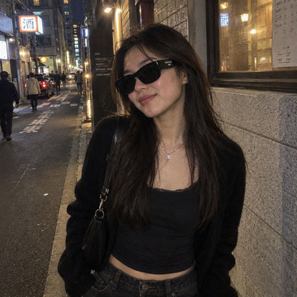 A woman wearing Balenciaga BB0390SK 001 Black Square Sunglasses, a black crop top, and a black cardigan stands on a city street at night.