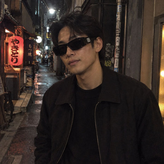 A man wearing Balenciaga BB0395S 001 Black Rectangular Sunglasseswith a small white logo and a black jacket, standing in a narrow, dimly lit street at night with Japanese lanterns visible in the background.