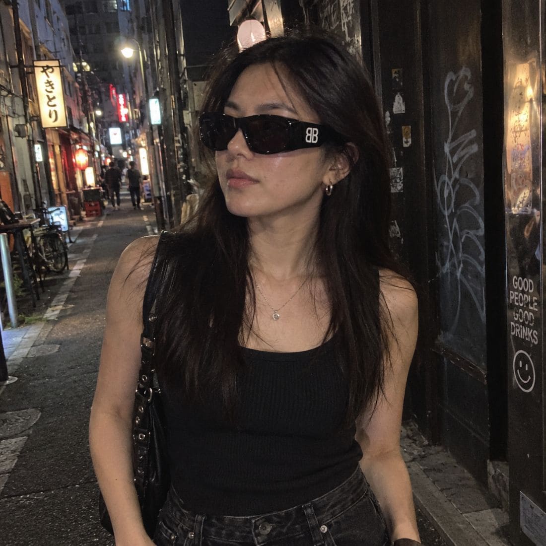 A woman wearing Balenciaga BB0395S 001 Black Rectangular Sunglasses with a white 'BB' logo on the temple, a black ribbed tank top, and dark jeans, standing on a city street at night.