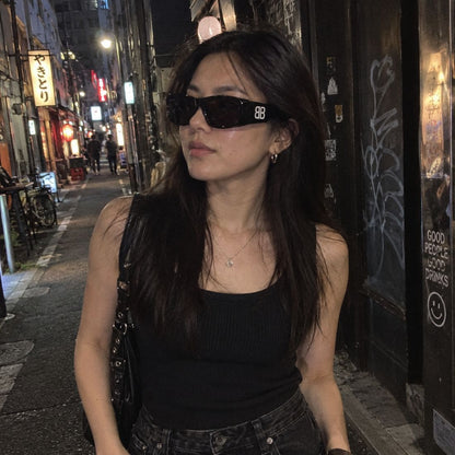 A woman wearing Balenciaga BB0395S 001 Black Rectangular Sunglasses with a white 'BB' logo on the temple, a black ribbed tank top, and dark jeans, standing on a city street at night.