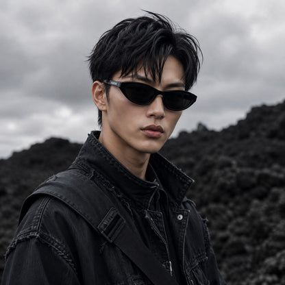 A man wearing Balenciaga BB0402S-001 Black Cat-Eye Sunglasses and a black distressed denim jacket, photographed outdoors against a dark, moody mountainous background under a cloudy sky.