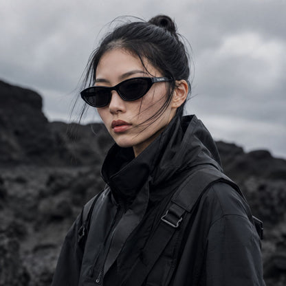 A woman wearing Balenciaga BB0402S-001 Black Cat-Eye Sunglasses and a black hooded jacket stands outdoors against a dark rocky landscape under an overcast sky.