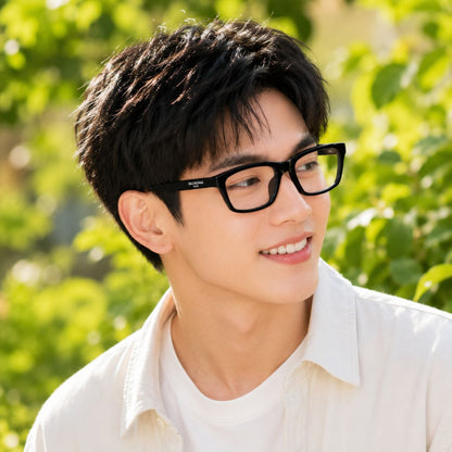 A smiling young man outdoors wearing Balenciaga BB0439O 001 Black Square Eyeglasses and a white collared shirt, with blurred green foliage in the background.