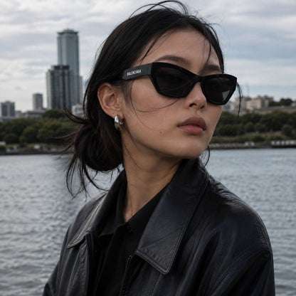 A woman wearing Balenciaga BB0447S Black Cat-Eye Sunglasses with a silver-tone frame detail and a black leather jacket, photographed outdoors with a city skyline and water in the background.
