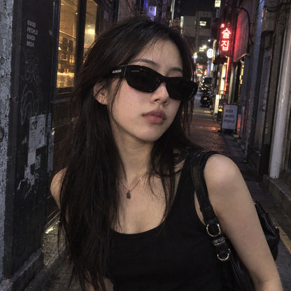 A woman wearing Balenciaga BB0448SK 001 Black Rectangular Sunglasses and a black tank top stands on a city street at night with blurred storefronts in the background.
