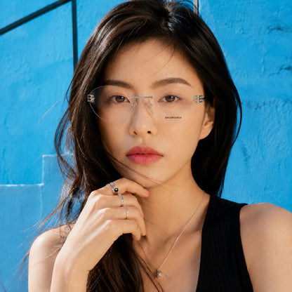 A woman with long dark hair wears Balenciaga BB0489O-001 Silver Rimless Butterfly Eyeglasses with a subtle metal bridge and small decorative side accents. She is posed against a bright blue textured wall, wearing a black sleeveless top and a delicate necklace.