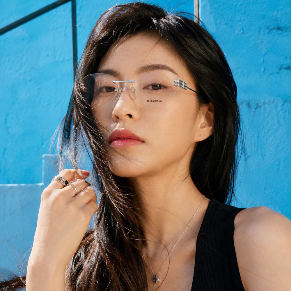 A woman with long dark hair wearing Balenciaga BB0489O-001 Silver Rimless Butterfly Eyeglasses with a silver bridge and small decorative corner embellishments, posed against a bright blue textured wall.