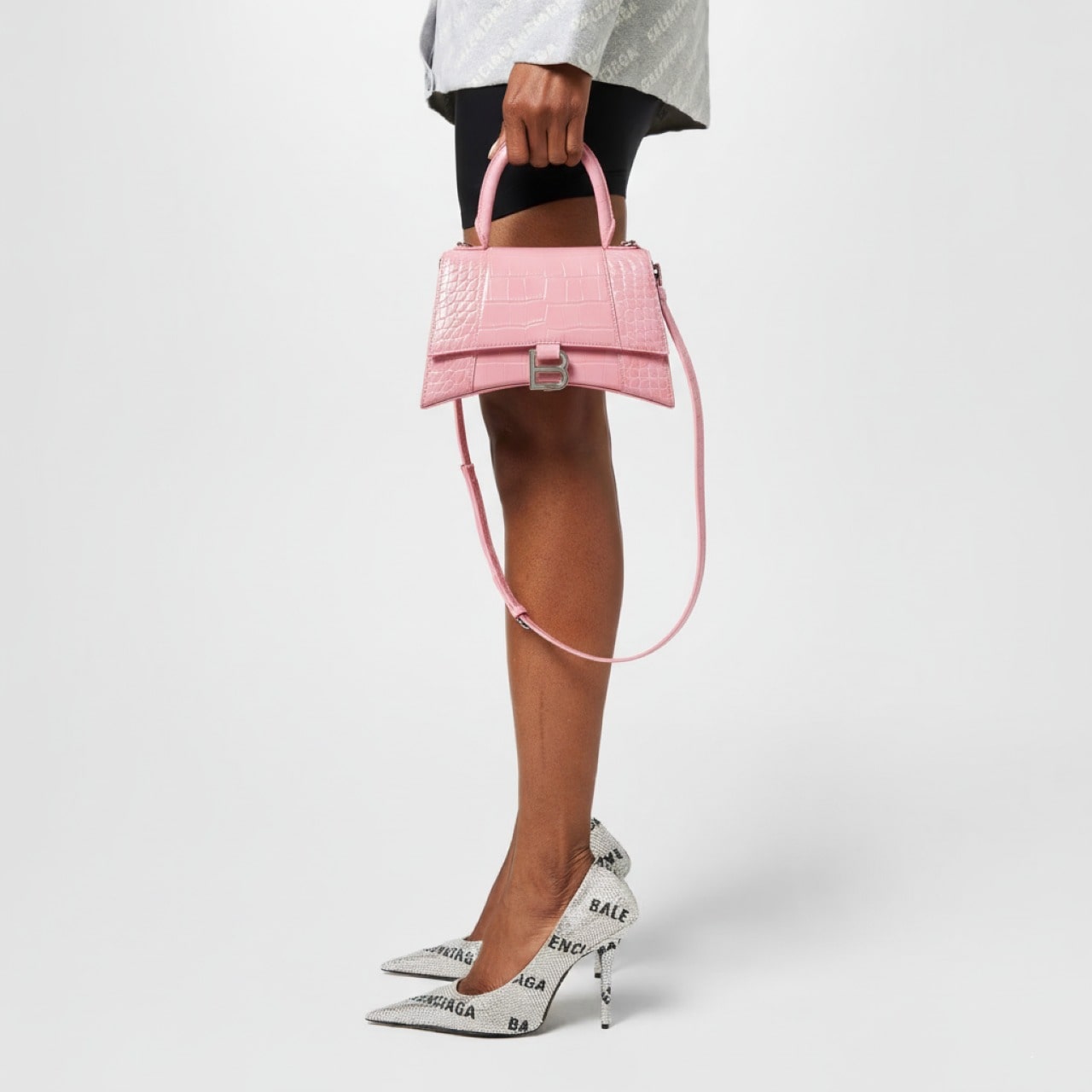 Person holding a Balenciaga Hourglass Small Handbag – Crocodile Embossed Pink Calfskin 5935461LR6Y5812 with white shoes featuring brand logos on a light gray background