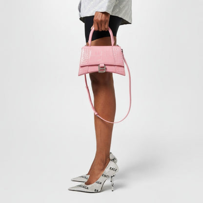 Person holding a Balenciaga Hourglass Small Handbag – Crocodile Embossed Pink Calfskin 5935461LR6Y5812 with white shoes featuring brand logos on a light gray background