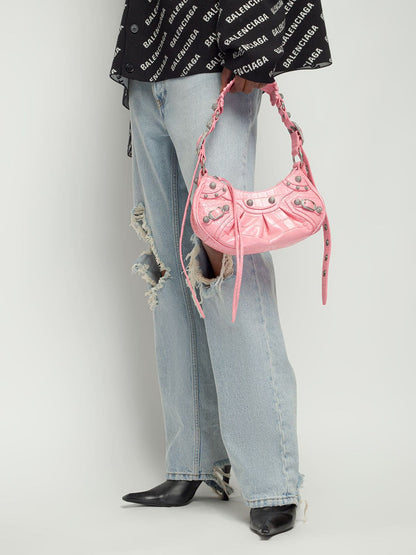 Person wearing a black Balenciaga top, light blue jeans, and holding aBalenciaga Le Cagole XS Shoulder Bag – Croc-Embossed Sweet Pink 67130923EJY5812 against a white background