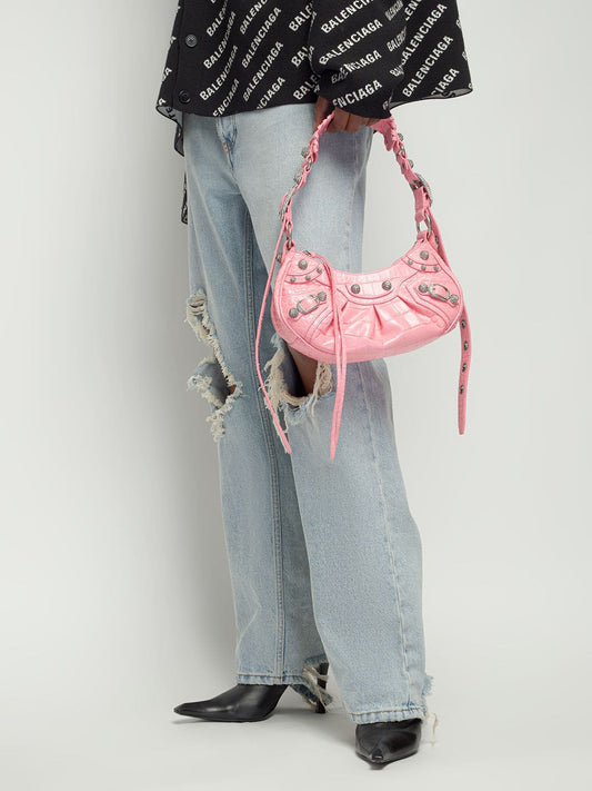 Person wearing a black Balenciaga top, light blue jeans, and holding aBalenciaga Le Cagole XS Shoulder Bag – Croc-Embossed Sweet Pink 67130923EJY5812 against a white background
