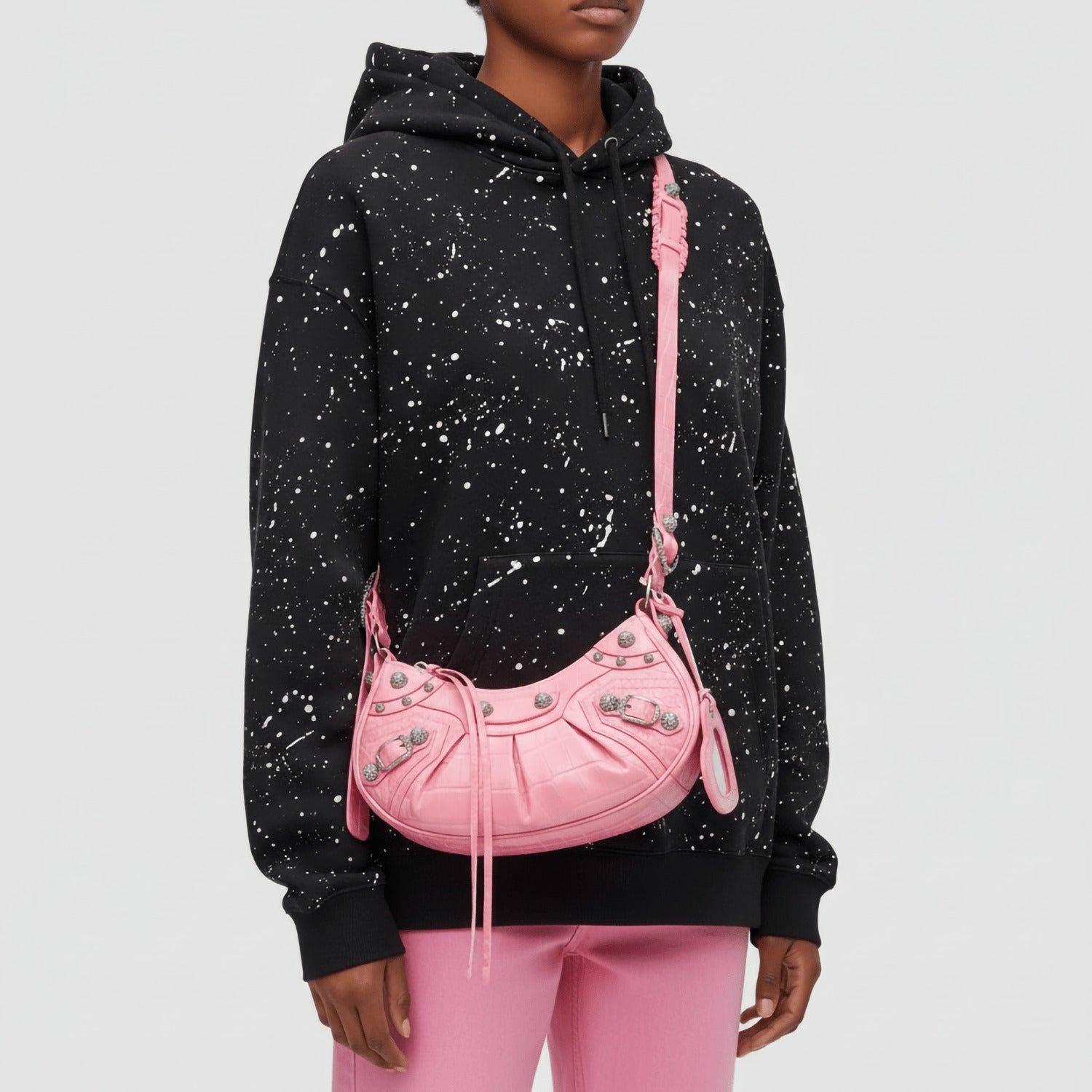Person wearing a black hoodie with white speckles and pink pants, holding a Balenciaga Le Cagole XS Shoulder Bag – Croc-Embossed Sweet Pink 67130923EJY5812.