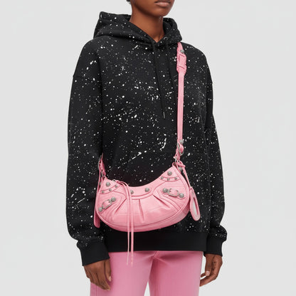 Person wearing a black hoodie with white speckles and pink pants, holding a Balenciaga Le Cagole XS Shoulder Bag – Croc-Embossed Sweet Pink 67130923EJY5812.