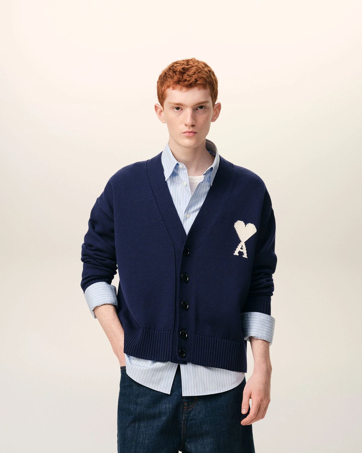Man wearing a Ami Paris Blue Wool Ami De Coeur Cardigan UKC380.KN0156.A116, light blue shirt, and dark jeans on a beige background