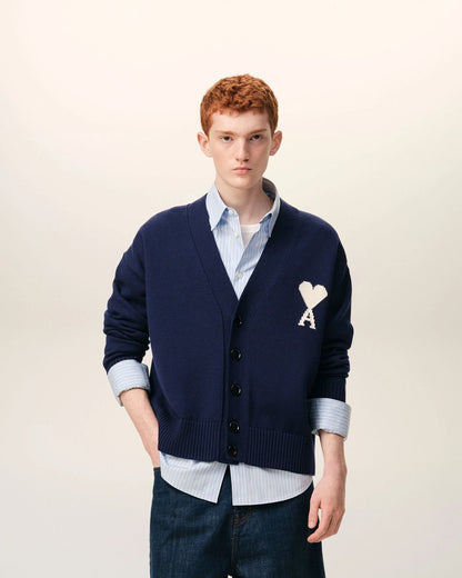 Man wearing a Ami Paris Blue Wool Ami De Coeur Cardigan UKC380.KN0156.A116, light blue shirt, and dark jeans on a beige background