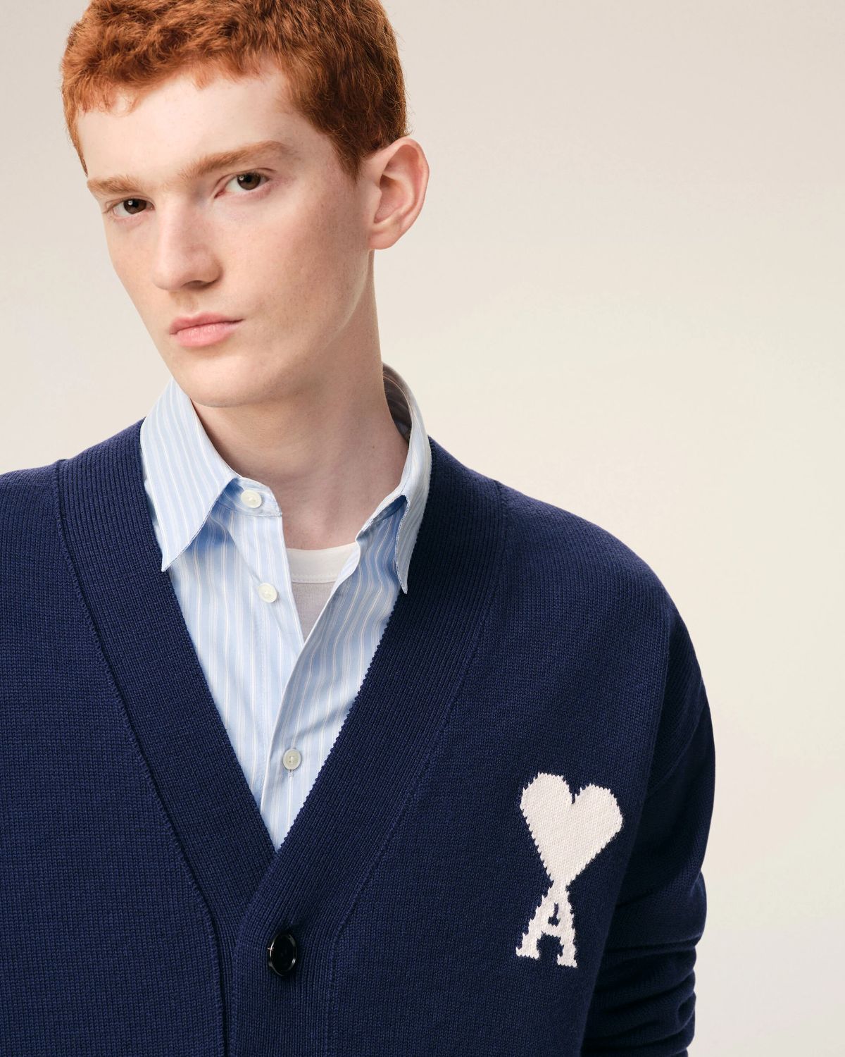 Man wearing a Ami Paris Blue Wool Ami De Coeur Cardigan UKC380.KN0156.A116 with a heart and 'A' emblem, light blue shirt underneath, on a beige background
